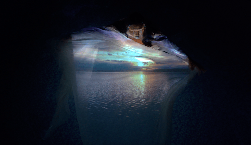 A figure swirling a large piece of white fabric in water. An image of a sunset is projected onto the fabric.