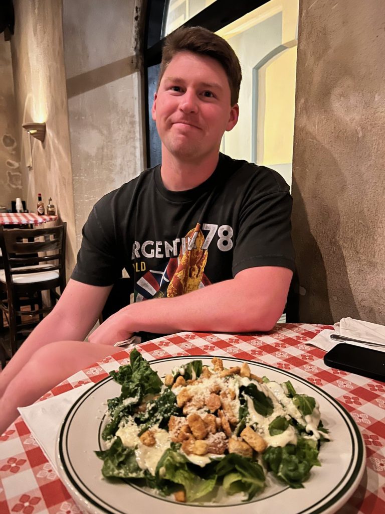 man smiling with a caesar salad in front of him on a red tablecloth. 