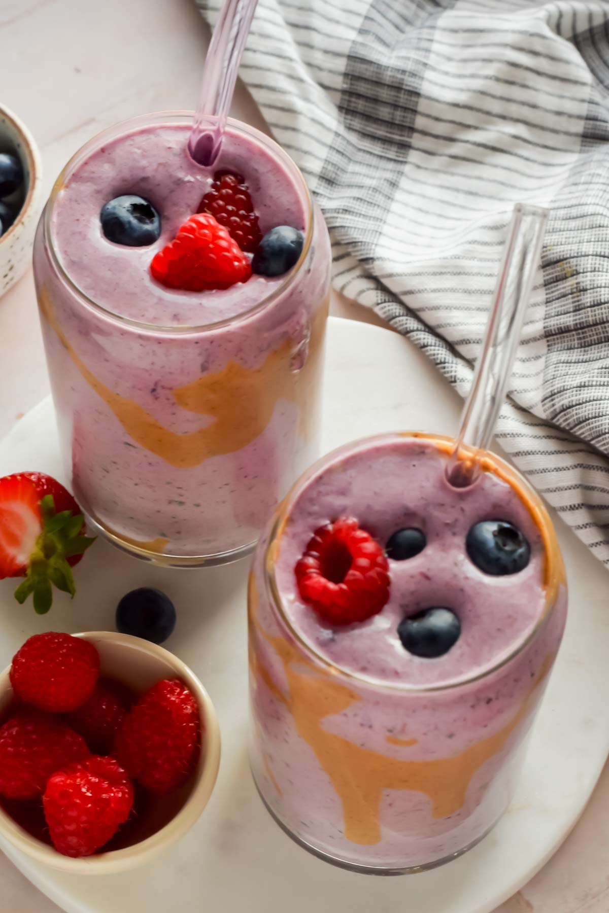 two berry smoothies in clear glasses with glass straws topped with fresh raspberries and blueberries.