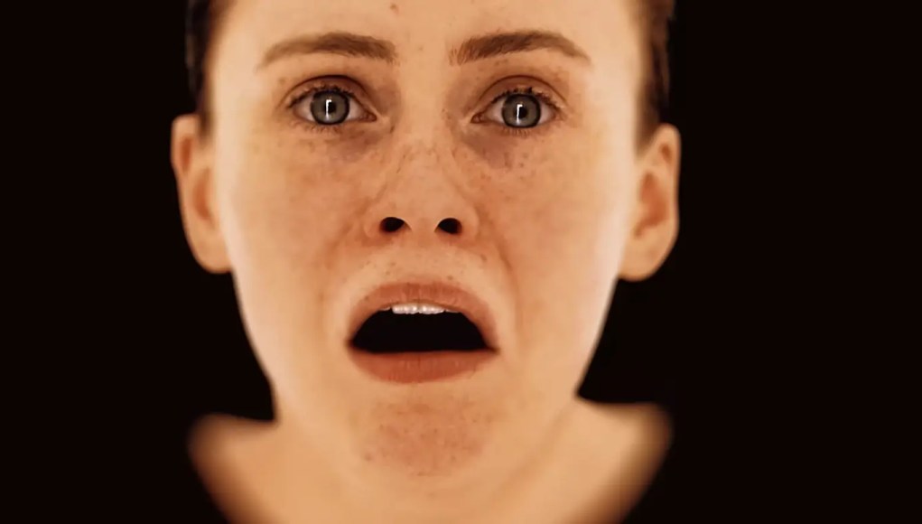 Primer plano de un rostro femenino digital generado por ordenador, con expresión de miedo o angustia, ojos muy abiertos y boca entreabierta sobre un fondo negro.