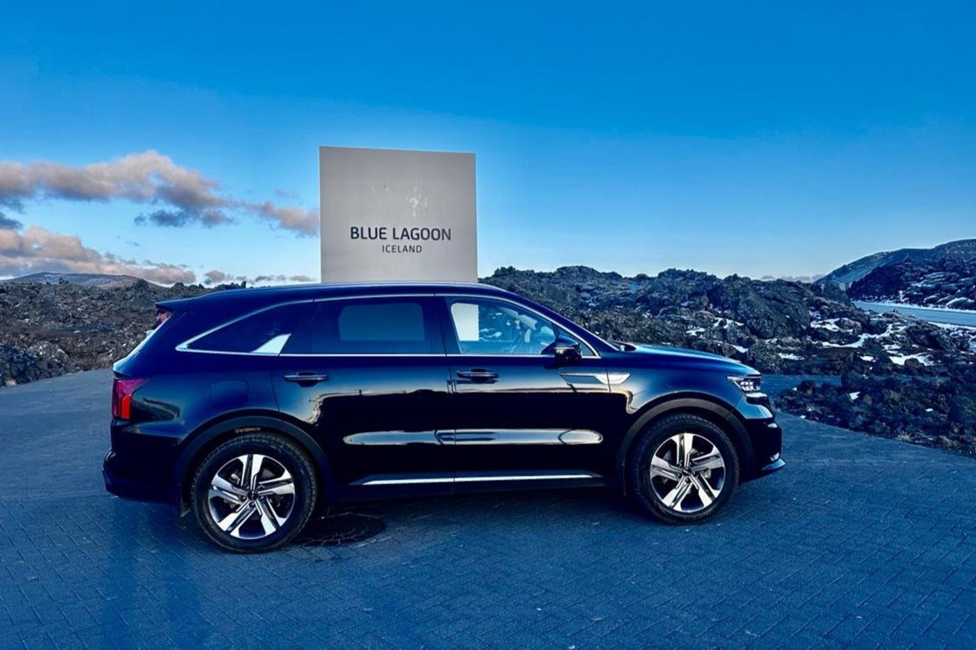 A black SUV is parked in front of a sign that reads "BLUE LAGOON ICELAND" with a rocky landscape and blue sky in the background.