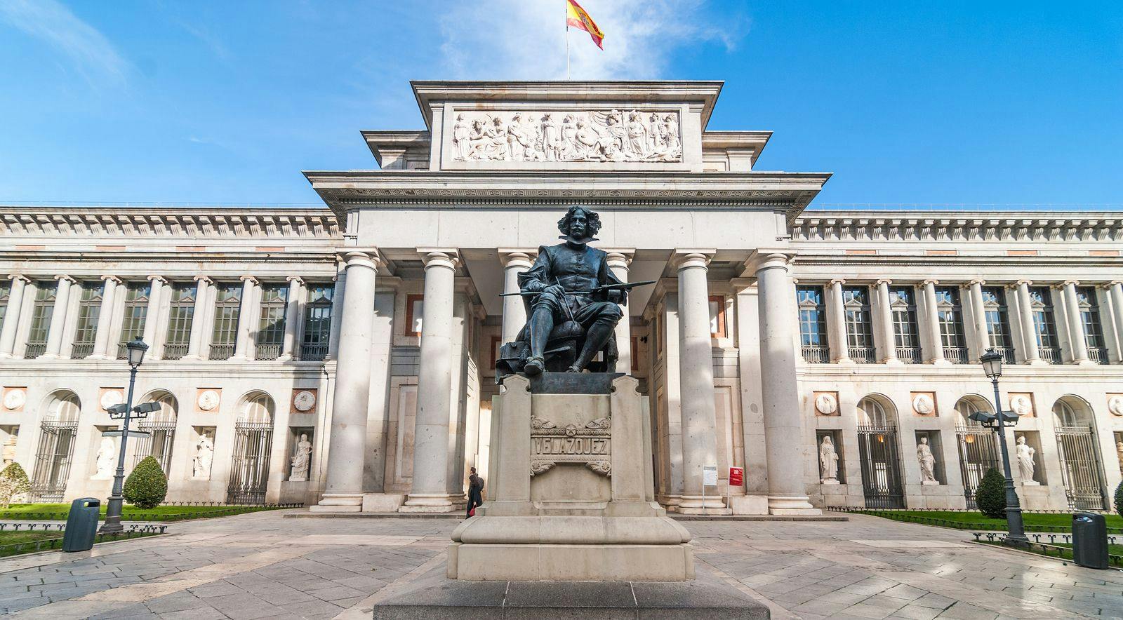 Statue of Diego Velázquez in front of the Prado Museum's columns, with a Spanish flag flying above.