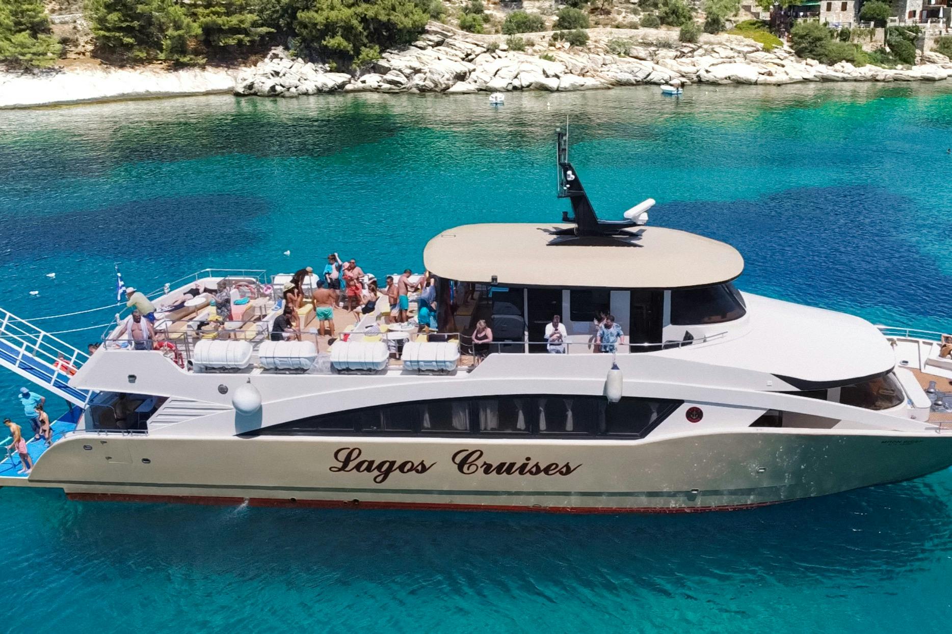 A cruise boat with "Lagos Cruises" written on the side, carrying people, is anchored in clear blue water near a rocky shoreline.