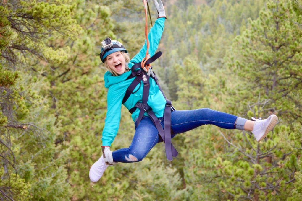 A person wearing a helmet and harness is ziplining through a forested area, smiling enthusiastically.