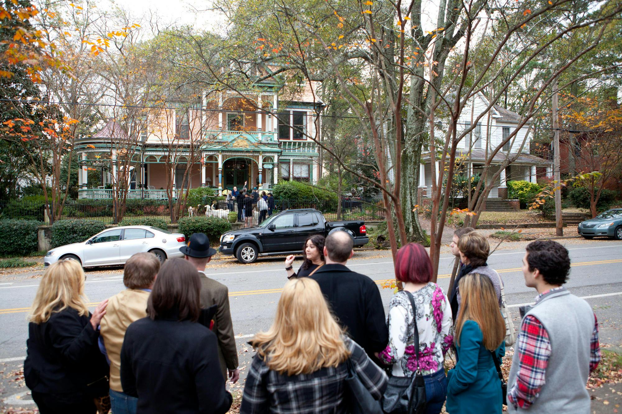 A group of people stand across the street from a large, ornate house surrounded by trees and parked vehicles.