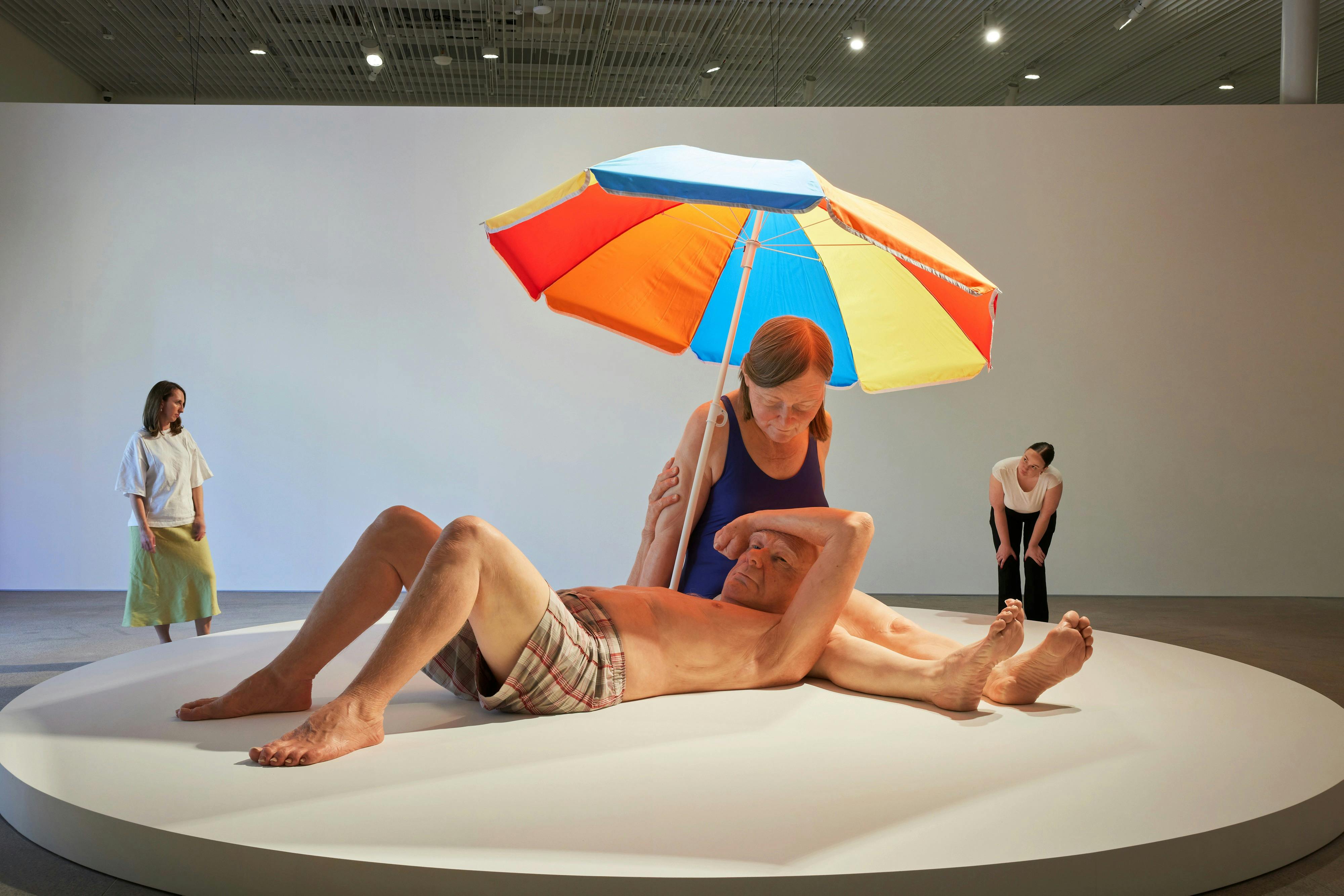 A sculpture of a man lying under a colorful umbrella with a woman seated behind him, in a gallery space with a viewer in the background.