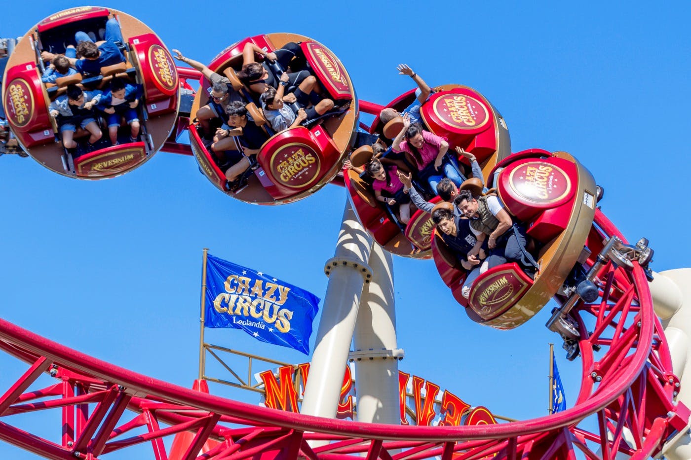 A group of people riding a spinning amusement ride in red seats, with a blue flag in the background under a clear sky.