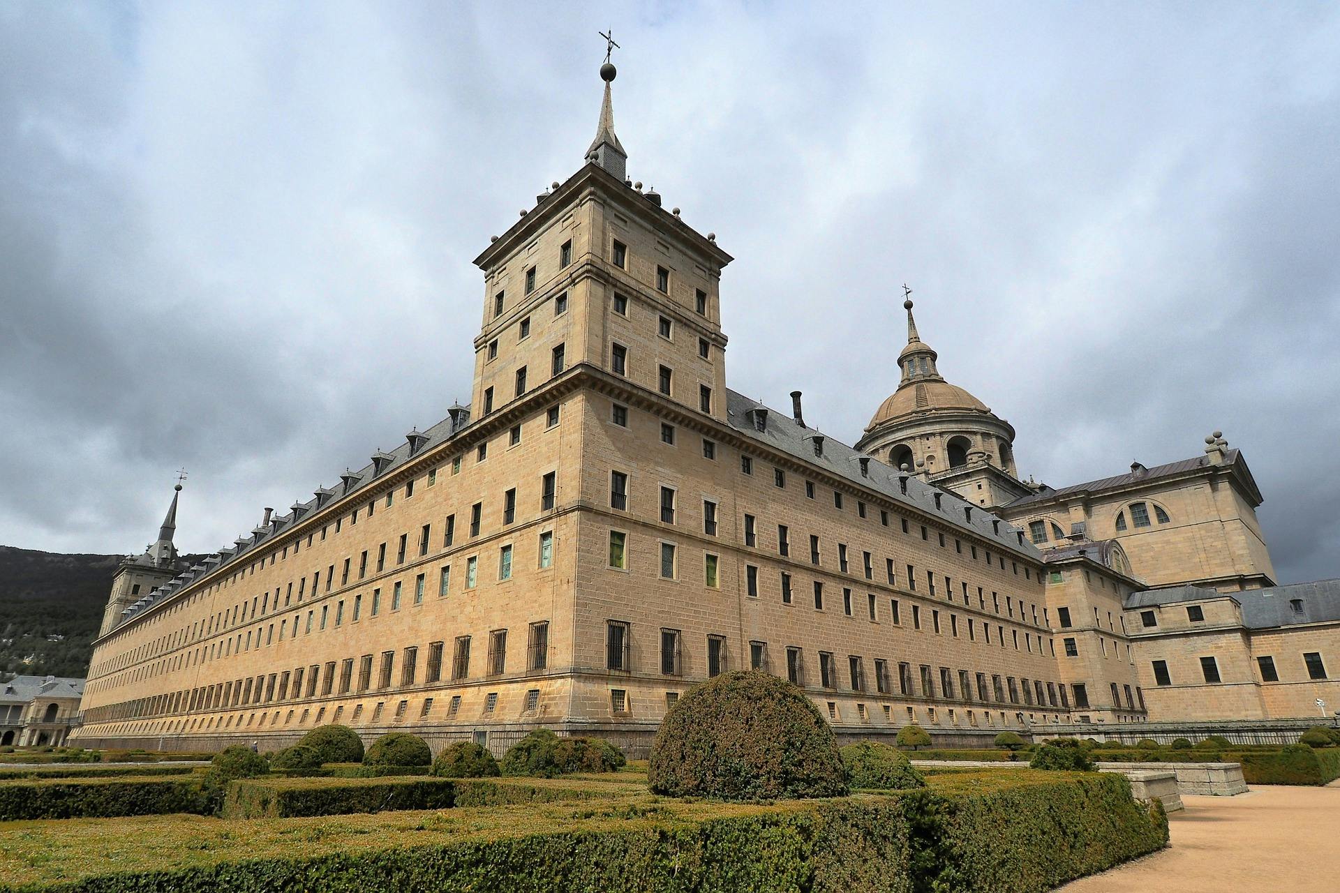 Large, historic stone building with a central tower, symmetrical windows, and a dome, surrounded by manicured hedges and a cloudy sky.