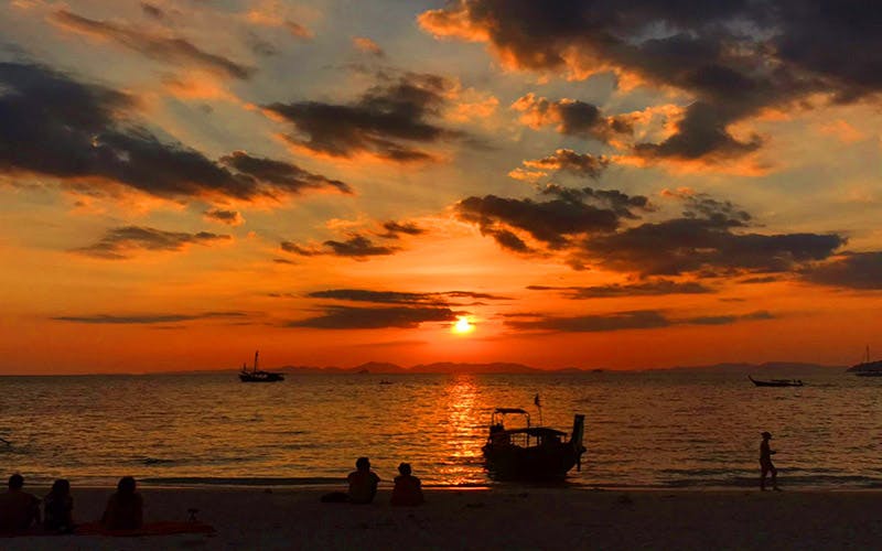 Two people sit on the beach, watching a sunset over the ocean with boats silhouetted against a colorful sky.