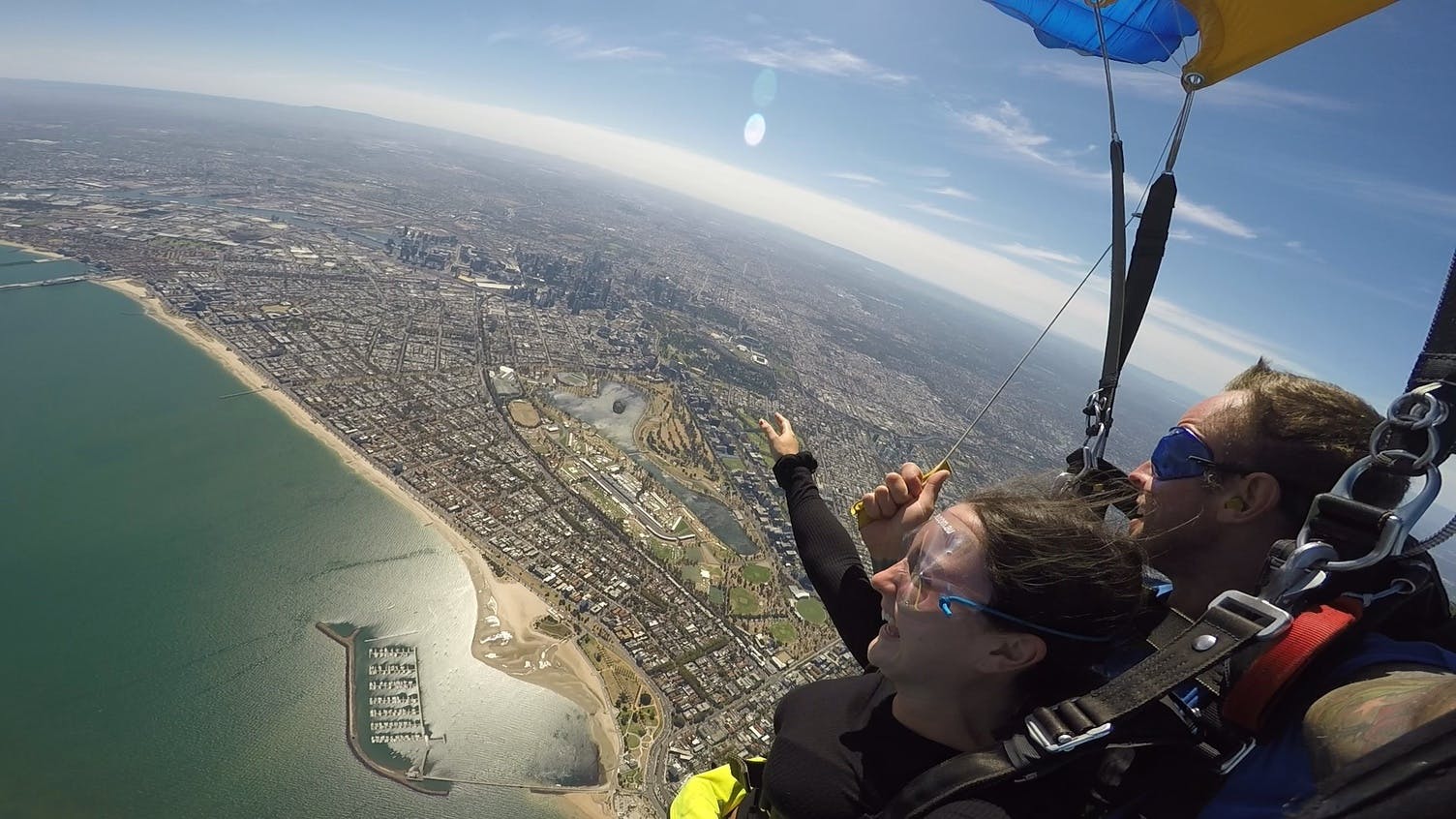 Person parachuting over a coastal city, pointing towards the ground, wearing a black outfit and goggles.