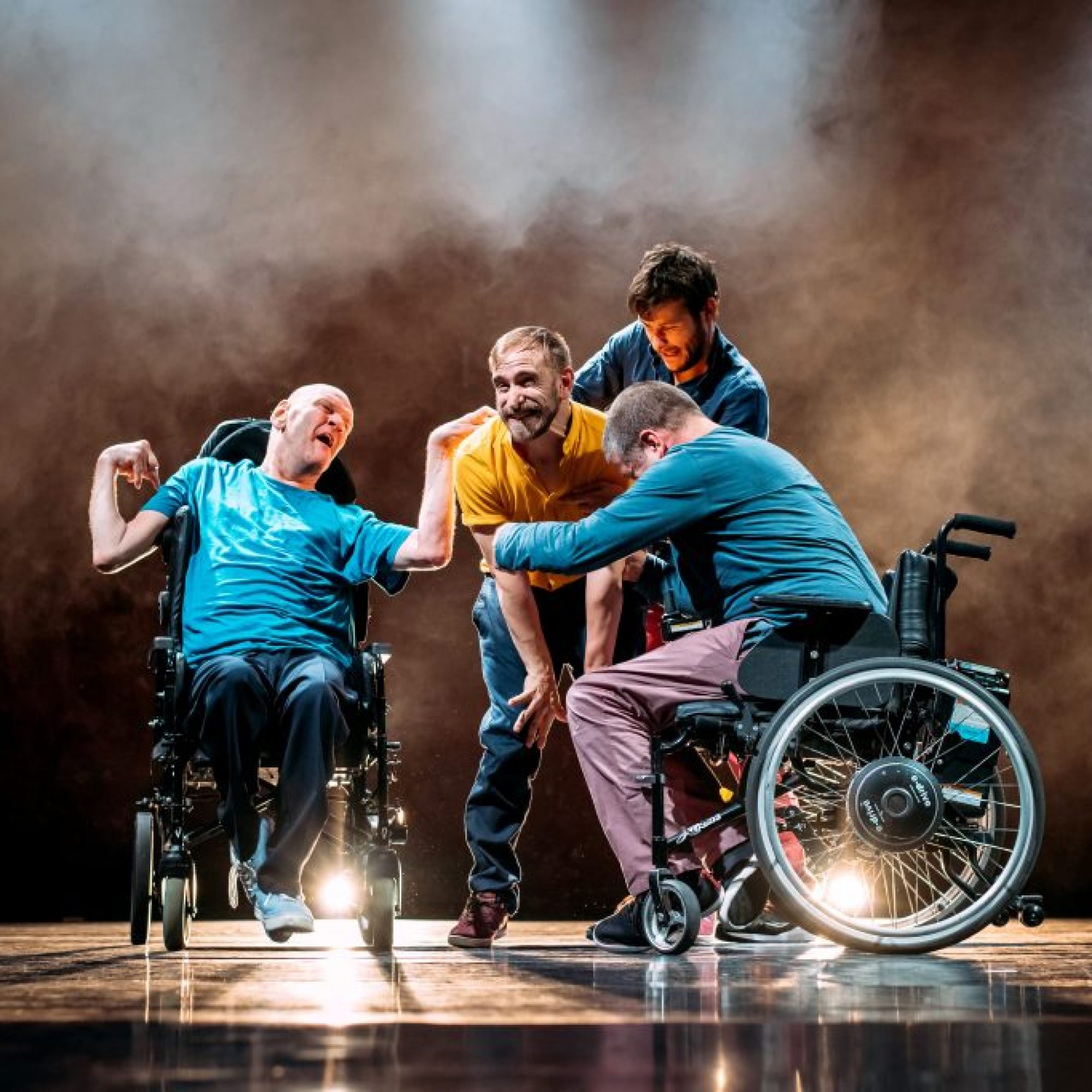 Four male performers clustered on a slightly smokey looking stage. Two of them are in wheelchairs, the other two are standing. Their ages appear to range from young to middle aged and they are dressed in various coloured shirts and jeans. They all appear to be reaching out to grab a man in a yellow T shirt who is standing in the middle of them all.