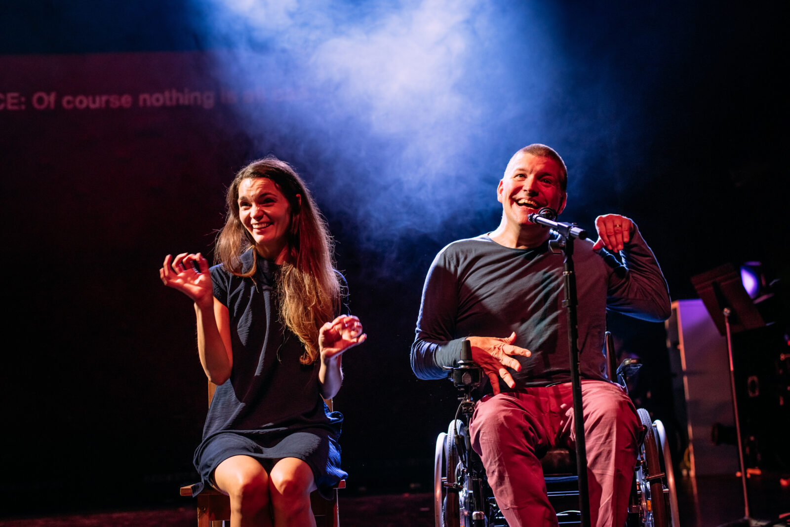 A man and a woman are sat next to each other on stage. They are both smiling. The man is talking into a microphone and the woman next to him appears to be signing what he is saying.