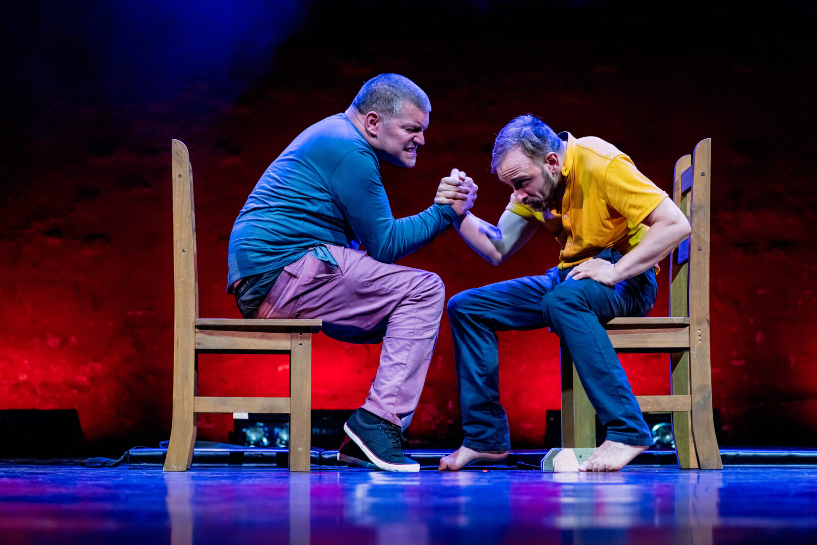 Two men sit opposite each other in wooden chairs. They are arm wrestling. Both men appear middle aged. The man on the left is wearing pink trousers and a blue long sleeved top and the man on the right is wearing a yellow shirt and blue trousers.