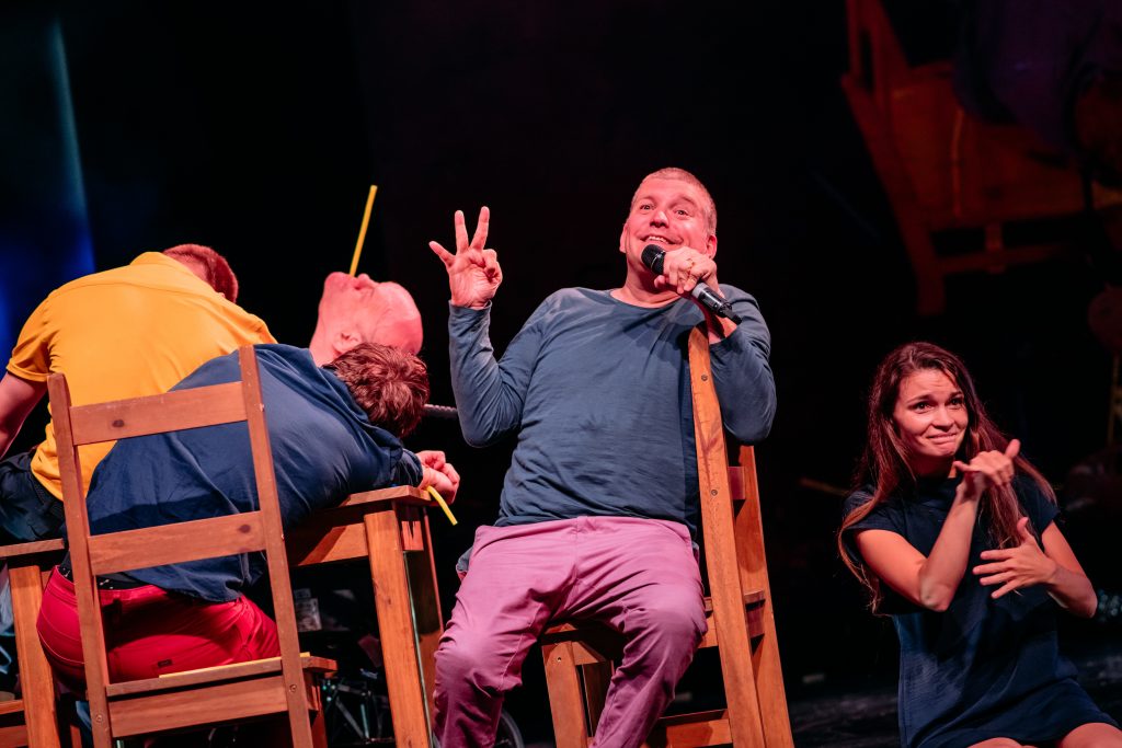 A smiling middle aged man sits on a wooden chair holding a microphone in one hand and flashing an 'OK' sign with his other hand. Beside him is a table and chairs that a few other men are sprawled out over them. On his other side there is a young woman with long hair sitting on the floor and signing.