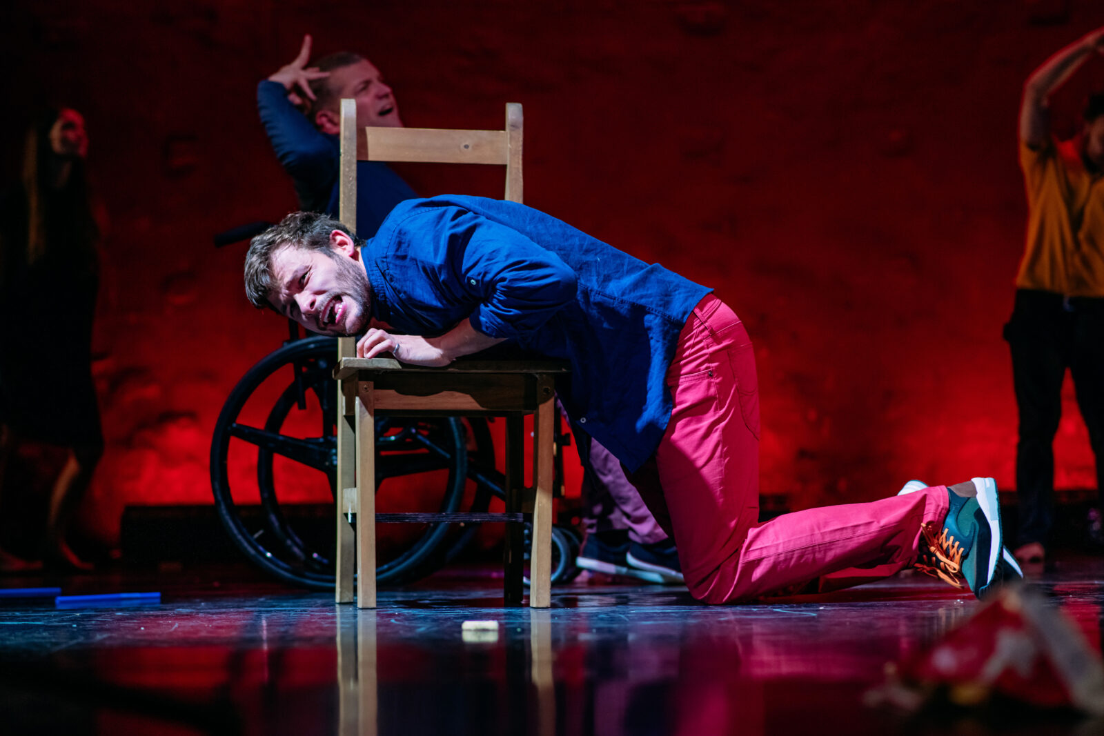 A young man with curly brown hair and a beard is on his knees on the ground, he is lying against a wooden chair, He is wearing pink trousers and a blue shirt. HIs expression looks upset.
