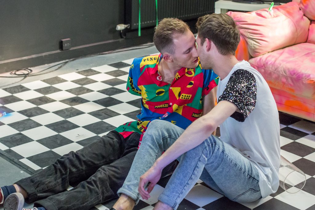 Two young men sitting on a black and white checkered floor are leaning into each other as if they are about to kiss. They both have brown hair, though the one on the right has darker hair than the other. The are both wearing skinny jeans. One is wearing a white t shirt with sequined sleeves, while the other is wearing a shirt with bright patterns on it.