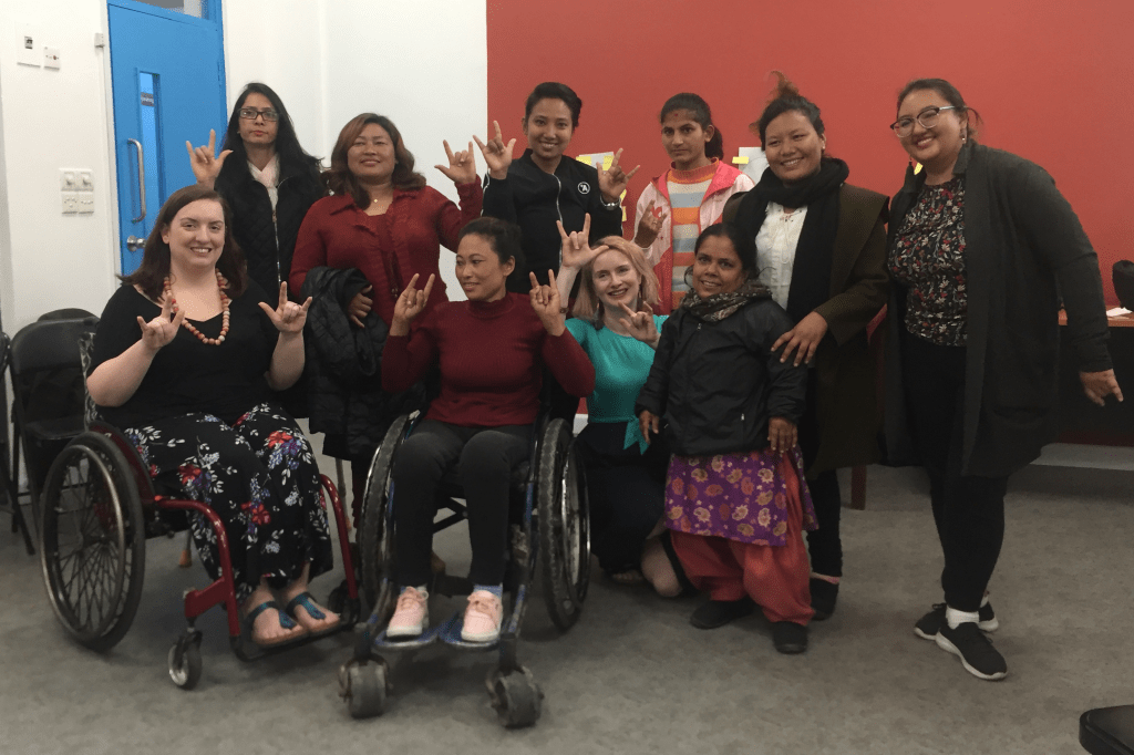 A large group of women pose for a photo. They all have their hands held up in signs, with their: thumbs, index fingers and little fingers held up and the other fingers held down.