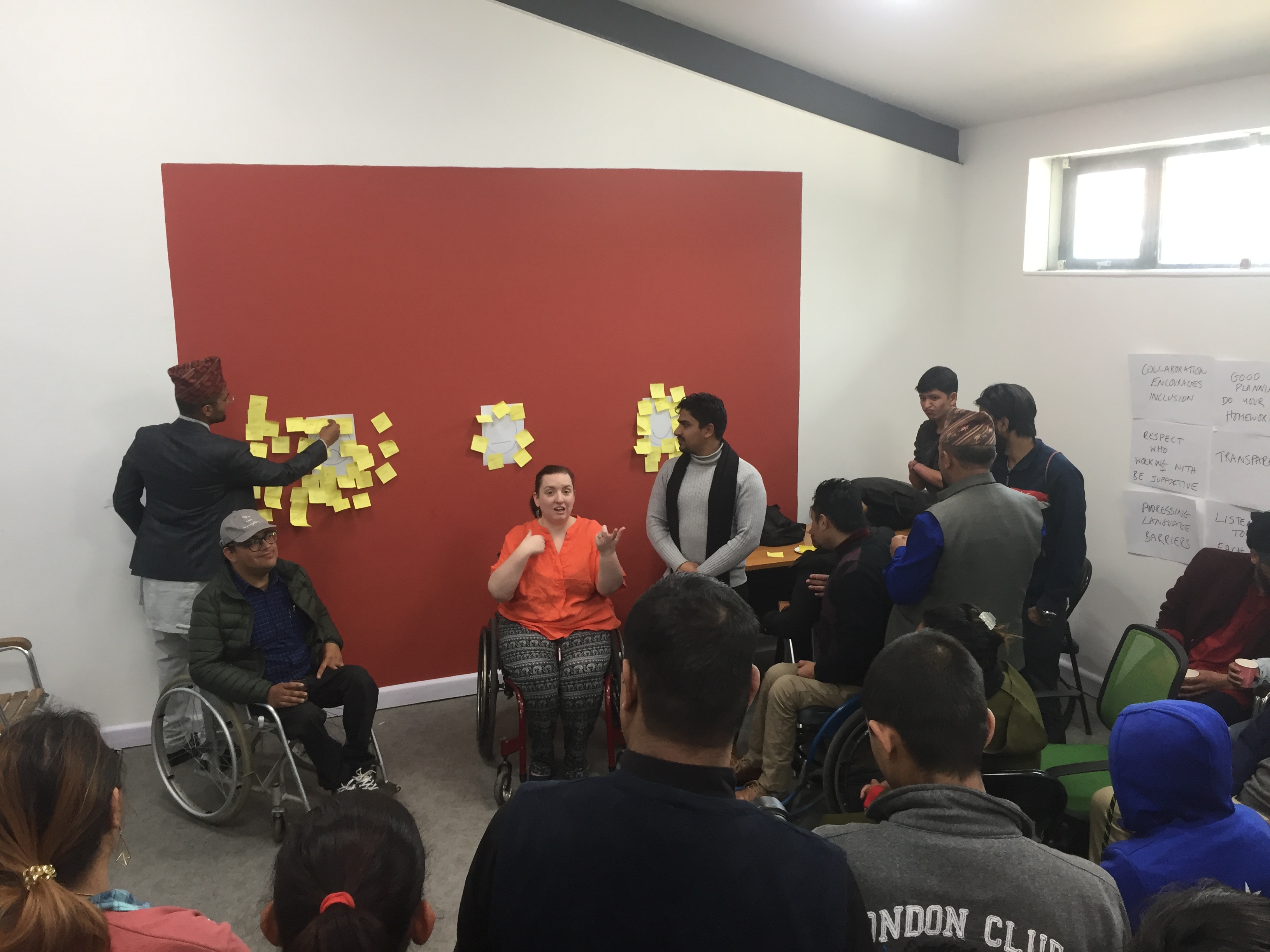 A room filled with people, some are sitting and some are standing. They are all facing a large red board with various clusters of sticky notes on it. In front of the board, there are two people in wheelchairs, a man and a woman. The woman is wearing an orange top and appears to be talking.