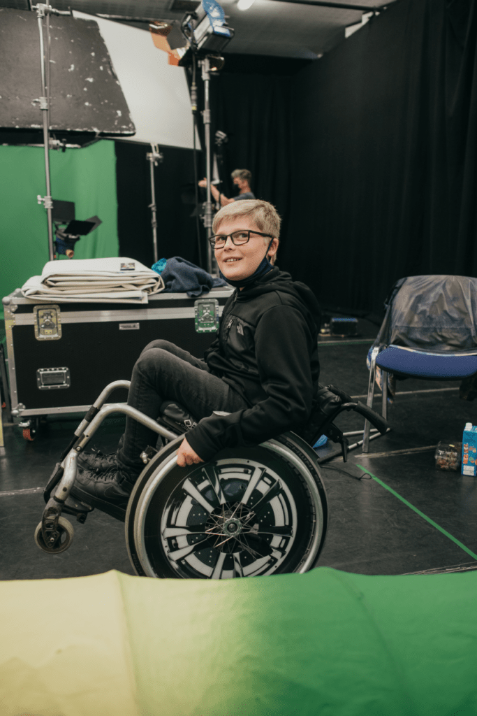 A photo of Ollie backstage. He is doing a wheelie on the large back wheels of his wheelchair with the small front two up in the air. He has a facemask pulled down at his chin