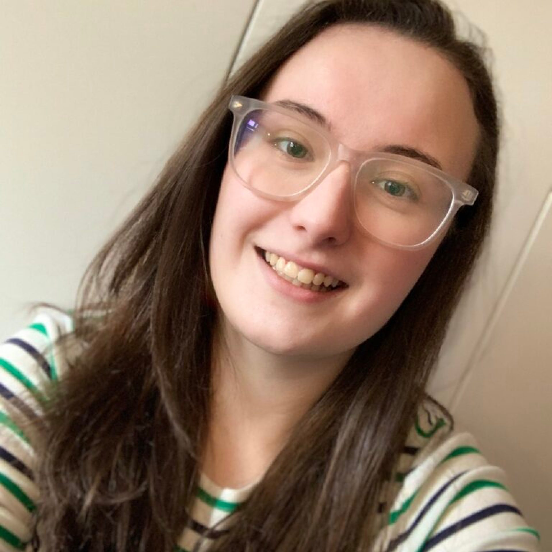 Photo of Rebekah. A young white wmoan in her 20s. They have long brown hair down to their chest and glasses with wide lenses and frames of clear plastic. They are smiling warmly at the camera.