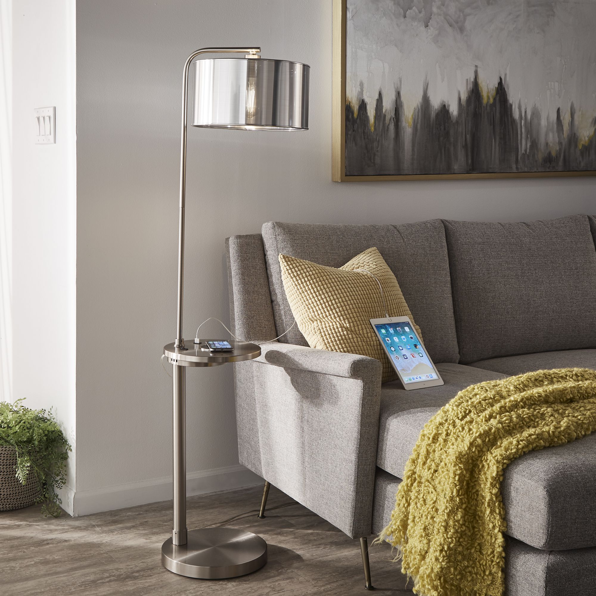This is the iNSPIRE Garrison Nickel Finish Metal Floor Lamp. It features a brushed nickel base and a drum lamp shade. A shelf is built into the middle of the base that also features a power outlet to charge electronics.