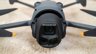Un dron gris oscuro con cuatro hélices cuenta con una cámara negra en su parte frontal; puedes ver la cámara de cerca mientras el dron se posa sobre una alfombra beige.