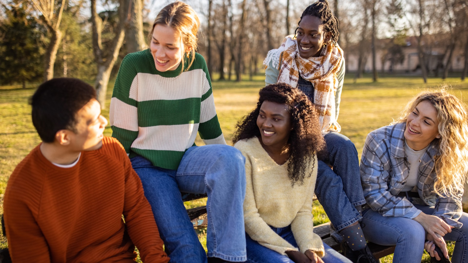 Group of five young people sat on a park bench smiling and talking with each other