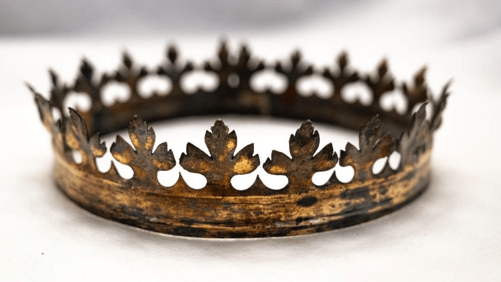 A tarnished gold crown ringed with fleur-de-lis type flowers sits on an off-white piece of fabric