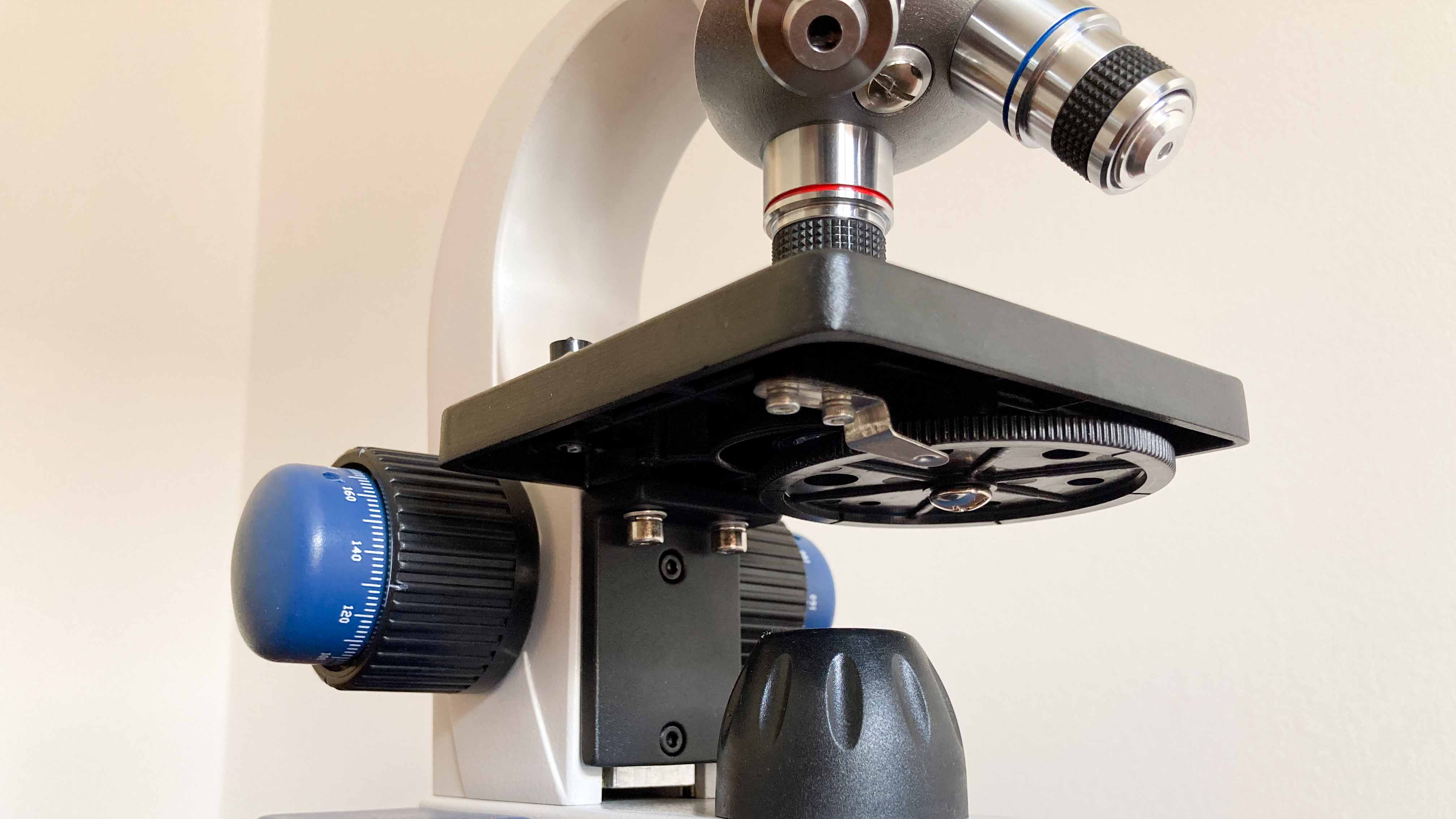 A close-up view displaying the microscope&rsquo;s single lens condenser, diaphragm, LED adjustment control, sturdy stage, coaxial coarse and fine focusing knobs and objective lenses