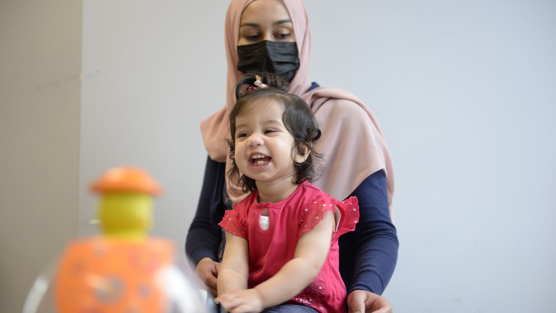 a smiling toddler wearing a red shirt sits in front of her mother who�s wearing a black medical mask and pink head scarf