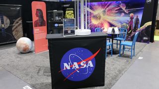 Una estantería negra con el logo de la albóndiga azul de la NASA se encuentra sobre una alfombra gris en el suelo de una convención de cómics.