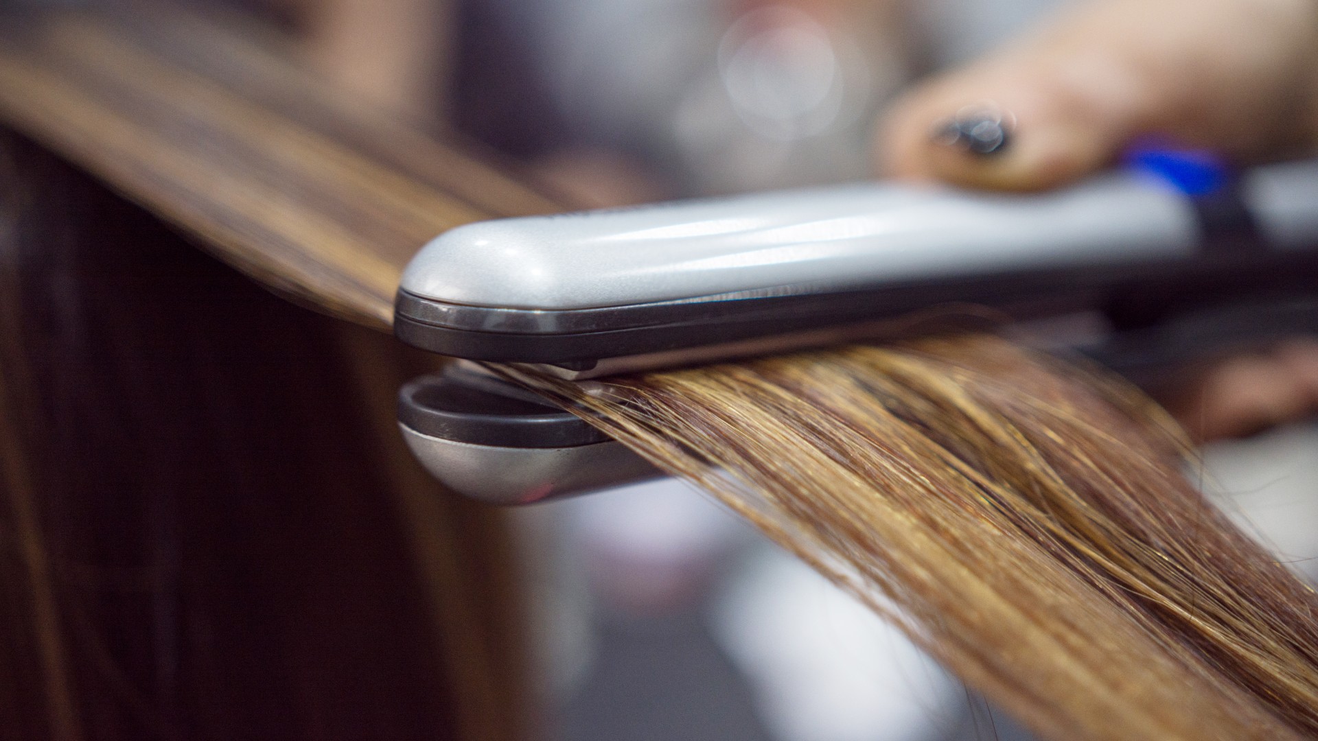 Close-up image of a pair of silver and black hair-straighteners being run through a woman&rsquo;s brown hair. Only the hair-straighteners are in focus in the image, the rest is blurred.