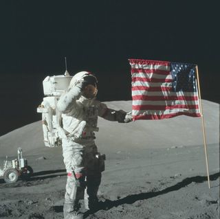 un astronauta con un voluminoso traje espacial blanco planta una bandera estadounidense en la polvorienta superficie gris de la luna