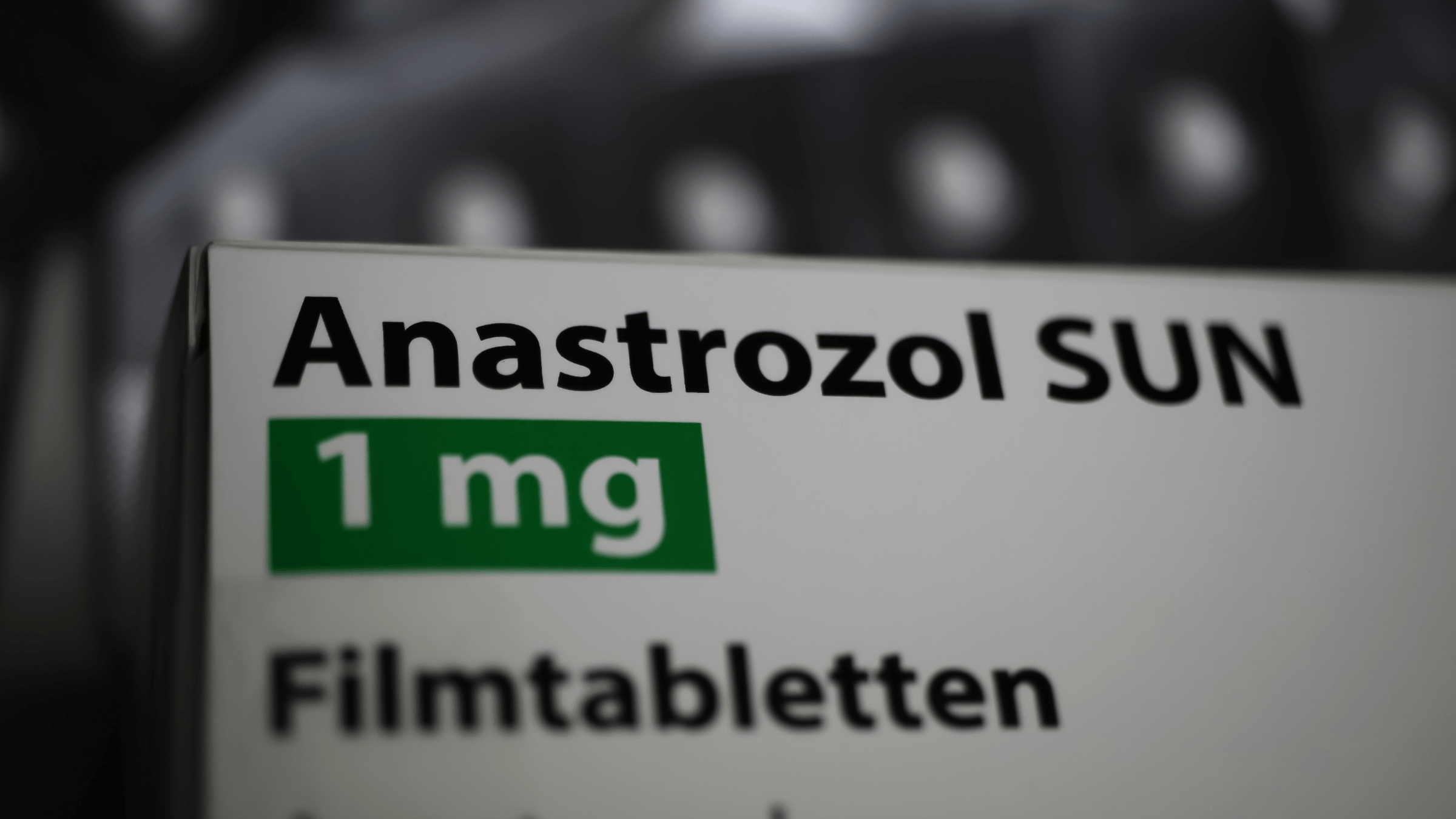 White box containing the drug anastrozol against a blurred, dark background. The drug name is written in black text on the box, as well as “1 mg” written in white text surrounded by a green box.