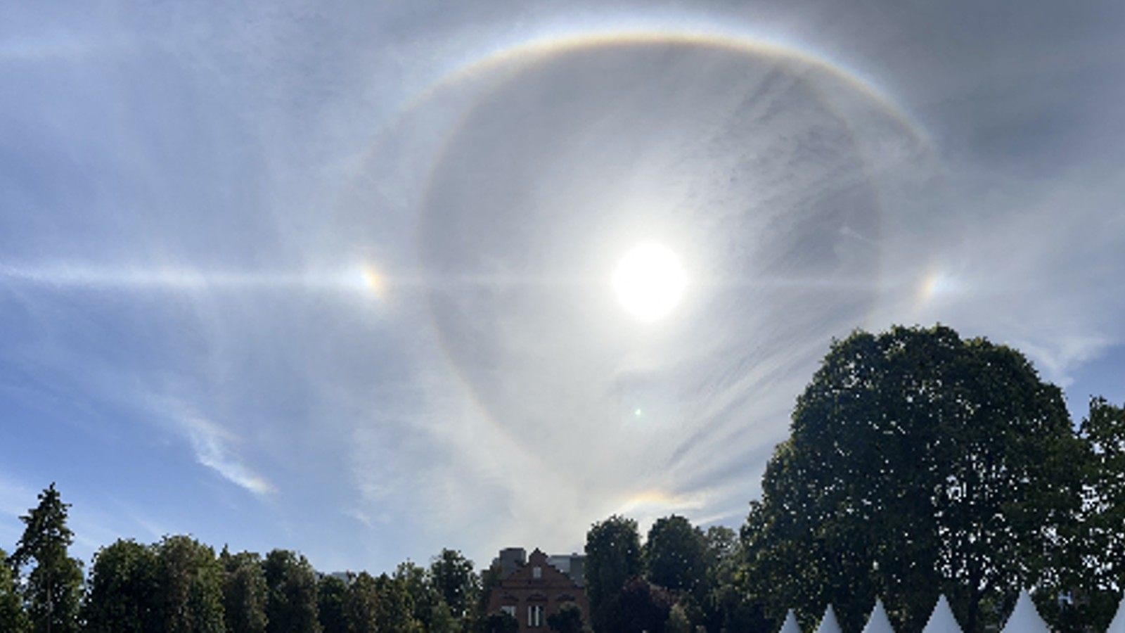 A blue sky with the sun surrounded by a white circle bisected with a white line and two bright spots either side of the circle.