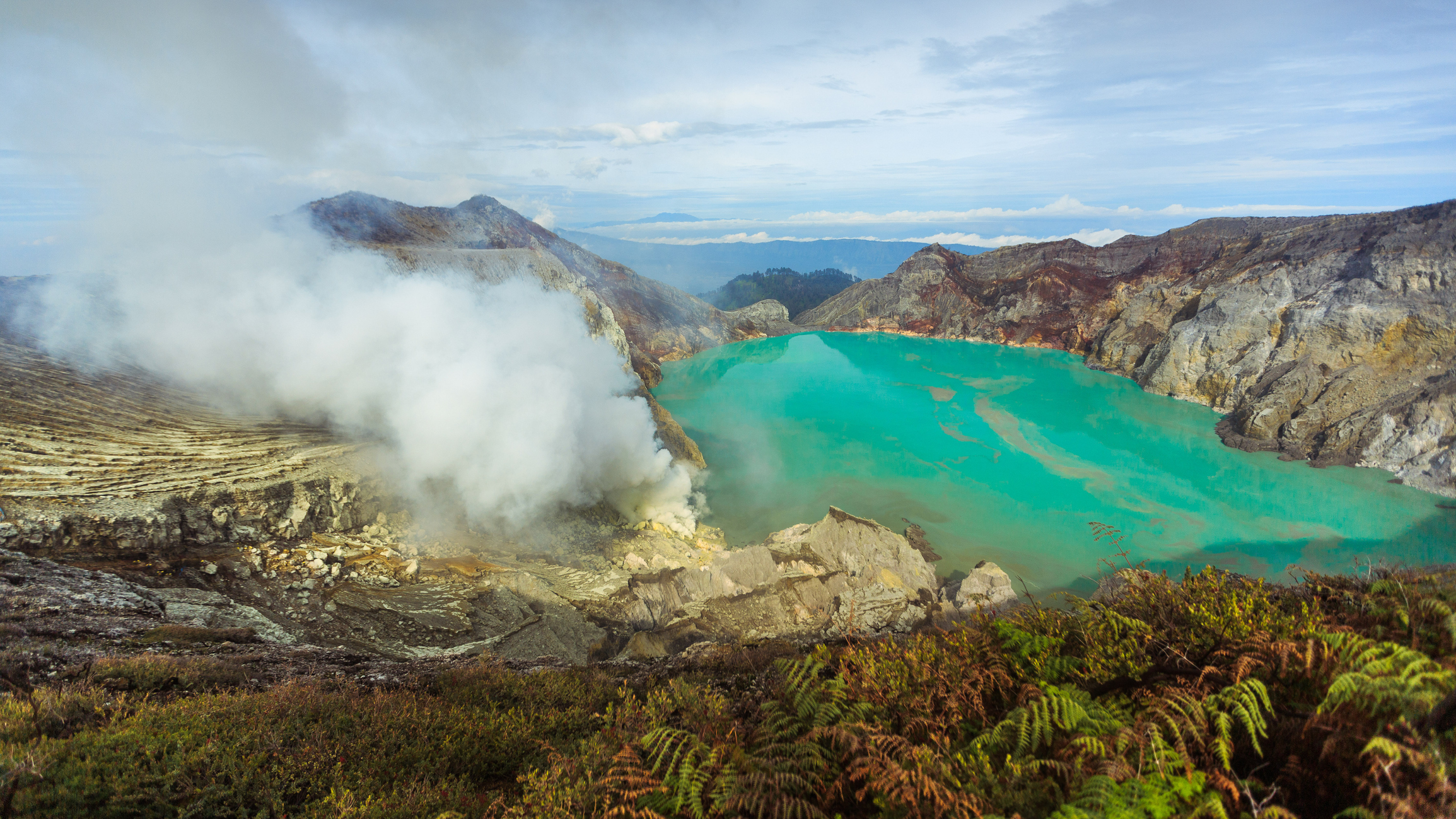 View of Kawah Ijen crater lake from the top of the volcano. The lake�s waters are bright turquoise and there is a plume of gas rising to the left.