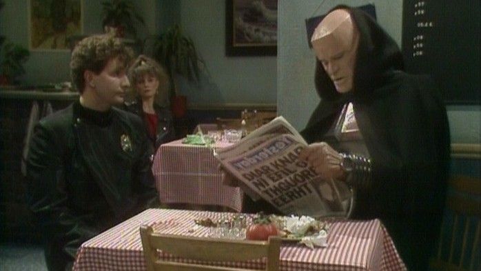 Fotograma de la comedia británica de ciencia ficción llamada Red Dwarf. Aquí vemos a Rimmer y Kryten en los episodios de 'Al revés'. Están sentados en un café leyendo un periódico al revés.