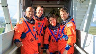 Cuatro astronautas con trajes espaciales de color naranja agrupados y de pie, sonriendo, en un brazo de acceso de lanzamiento utilizado para lanzamientos de cohetes.