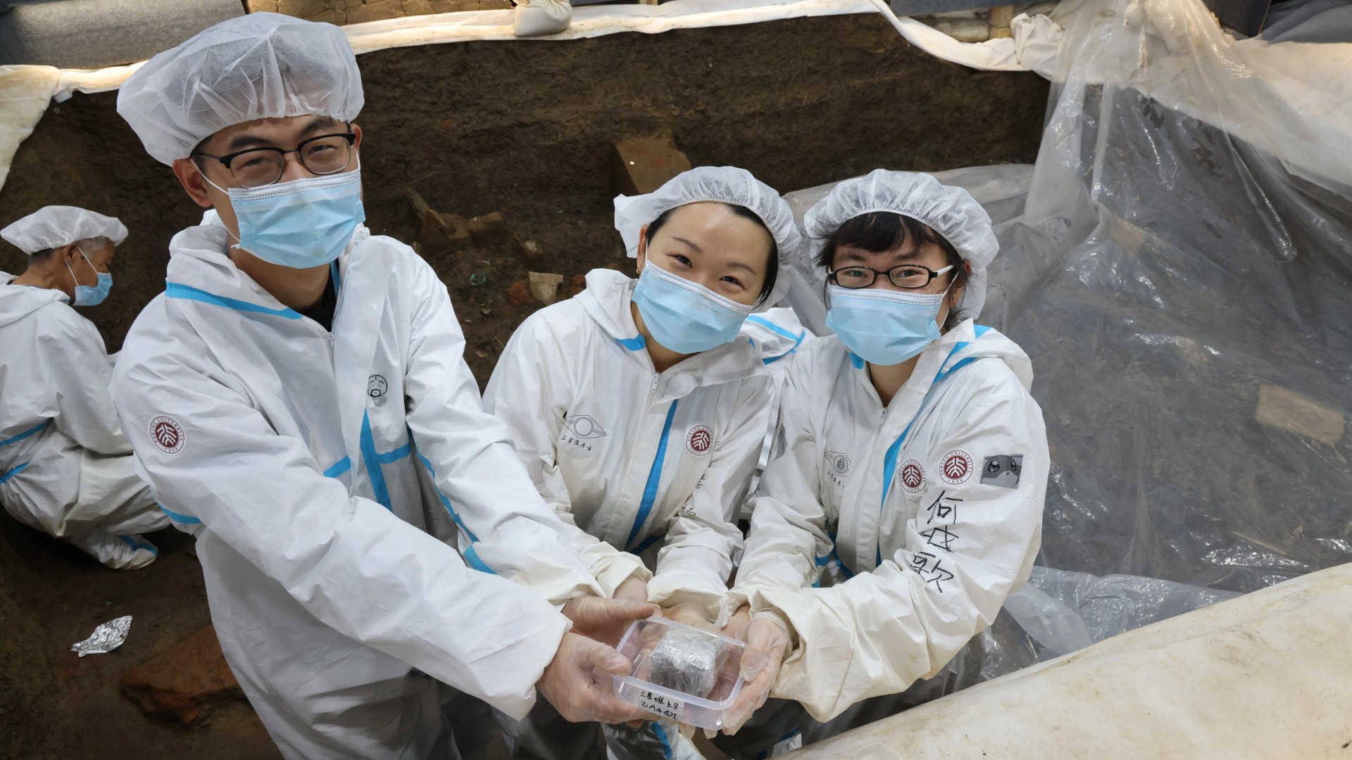 Archaeologist Li Nan (center) with two members of Peking University&rsquo;s archaeological team at the Sanxingdui archaeological site in China&rsquo;s Sichuan province. (plus one more person working in the back). Everyone is wearing hair covers, face masks, and white protective suits.