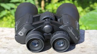 Prismáticos EclipSmart 12x50 colocados sobre una superficie de madera con follaje verde detrás