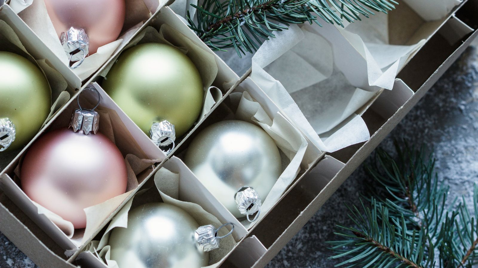 A box of pastel colored baubles with cardboard dividers between them. Pieces of fir tree cut up and laying around the box.