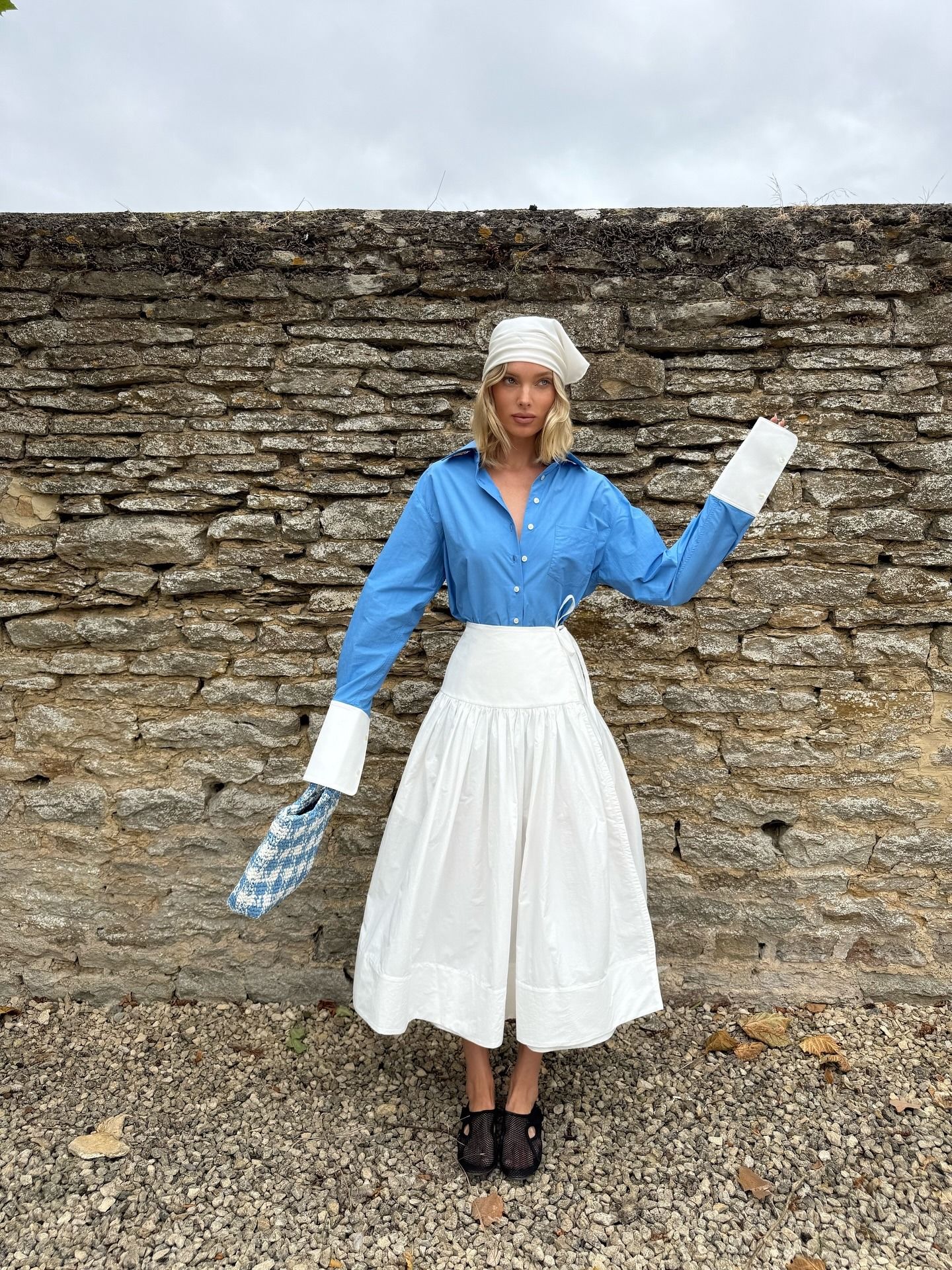 Elsa Hosk wearing a head scarf, Helsa blue button-down shirt and white poplin midi skirt, and Alaia fishnet slippers in the Cotswolds