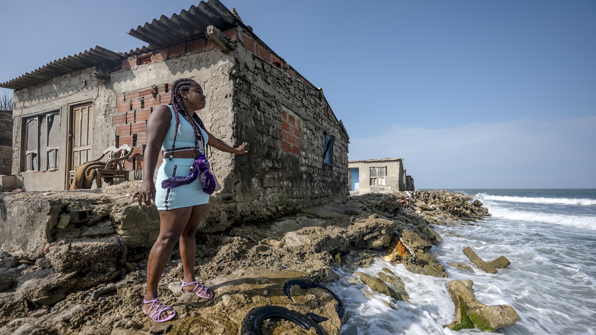 A woman stands by a weathered house, with the ocean only feet away.