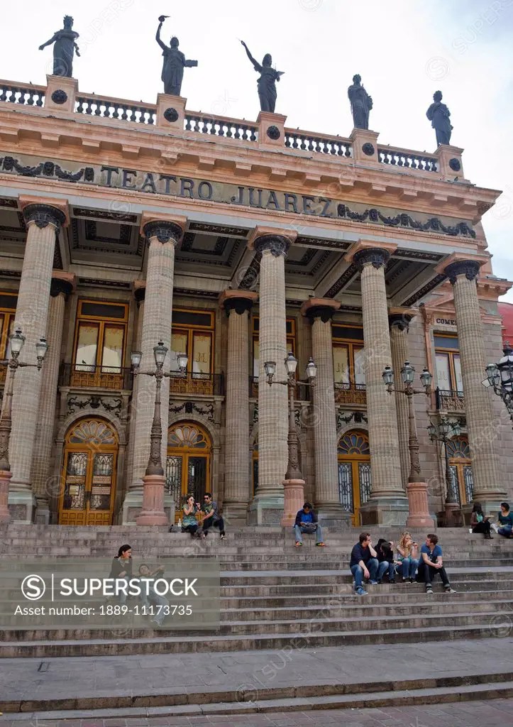 People sitting on the steps of an old spanish theatre with pillars and