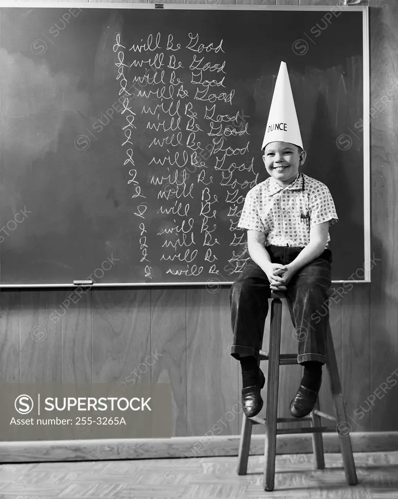 Boy wearing a dunce cap sitting in front of a blackboard SuperStock