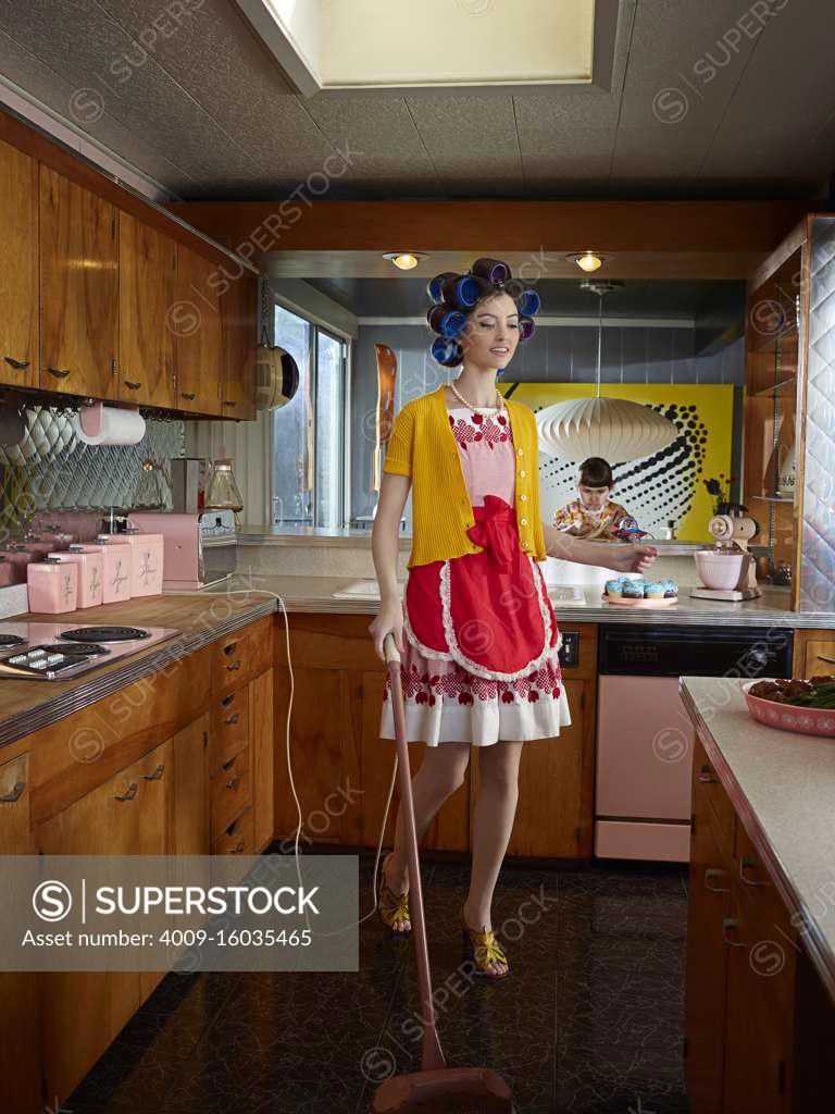 Woman vacuuming kitchen while wearing hair rollers in a midcentury