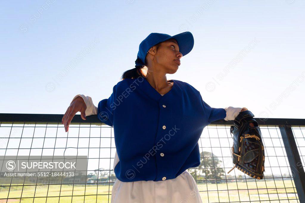 Mixed race female baseball player wearing glove, leaning on fence in