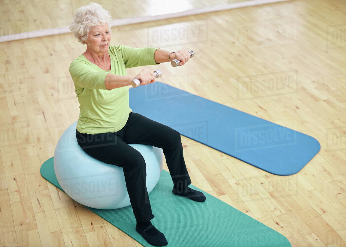 Senior female sitting on a fitness ball and lifting dumbbells. Old