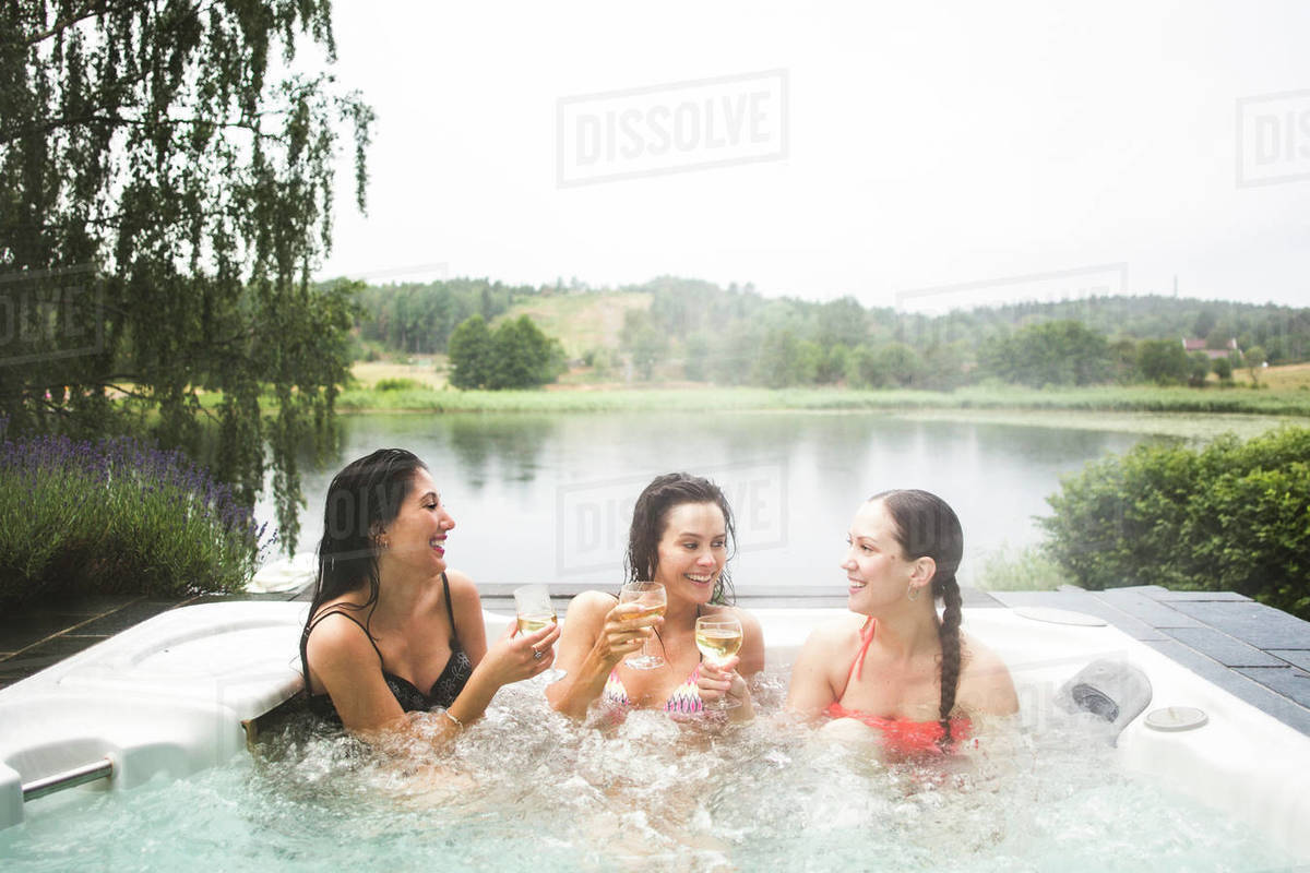 Cheerful female friends enjoying wine in hot tub against lake during