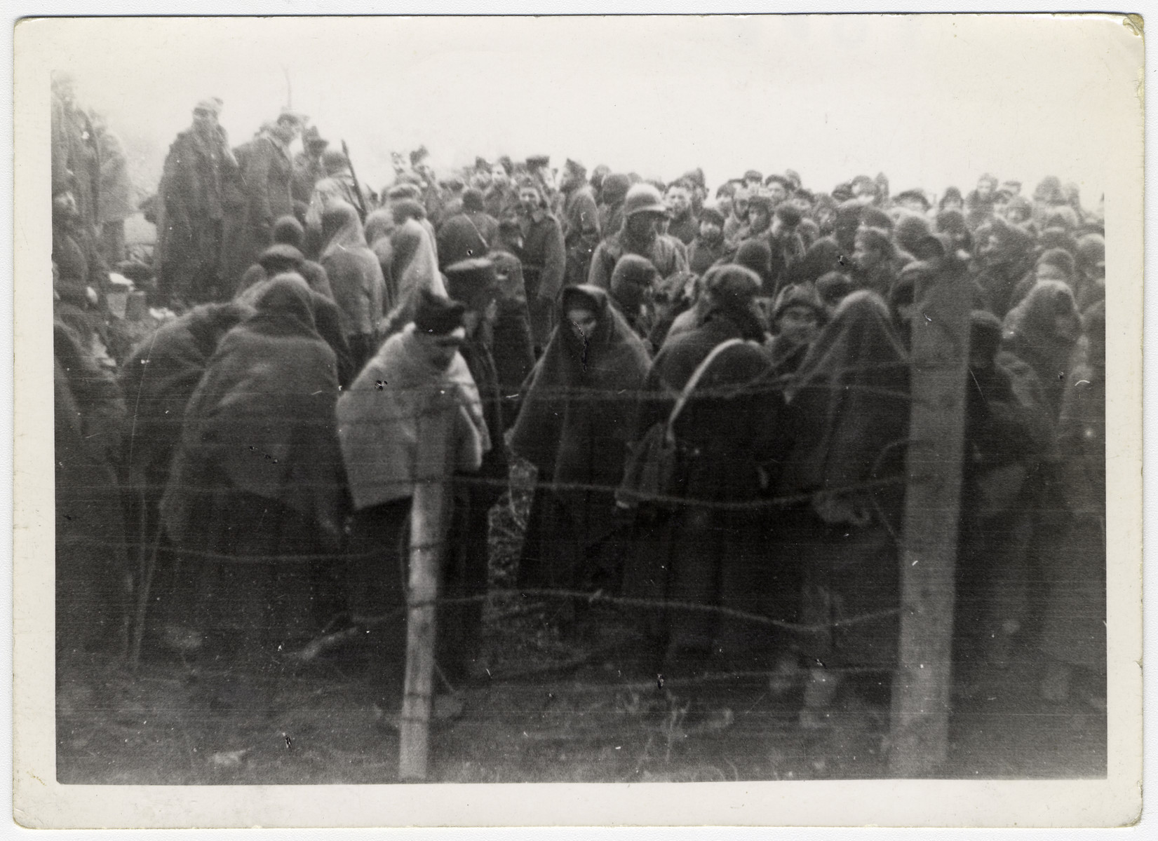 Survivors wearing blankets for warmth crowd together behind a barbed